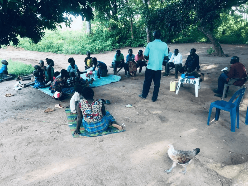 A pastor speaking to a group gathered outside under trees.